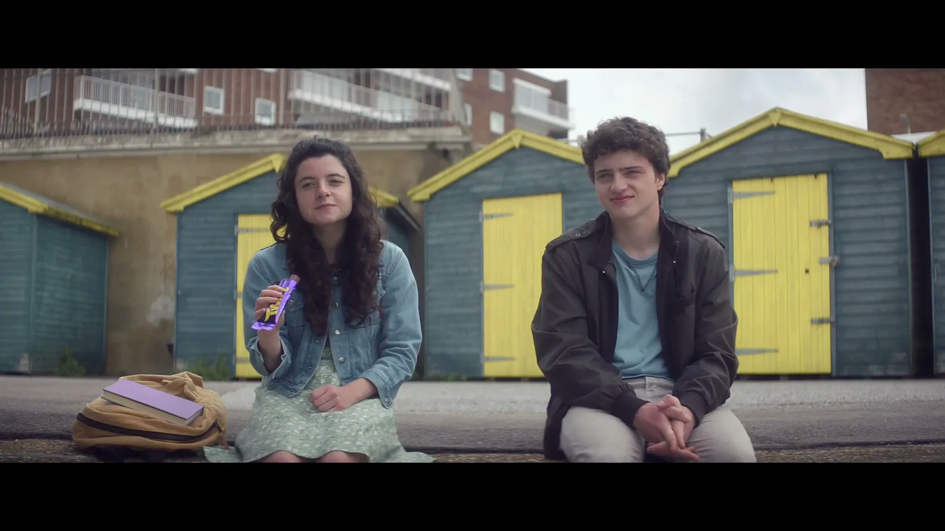 A young woman and man sit on a concrete ledge, with the woman holding a Cadbury chocolate bar, against a backdrop of colorful beach huts and buildings.