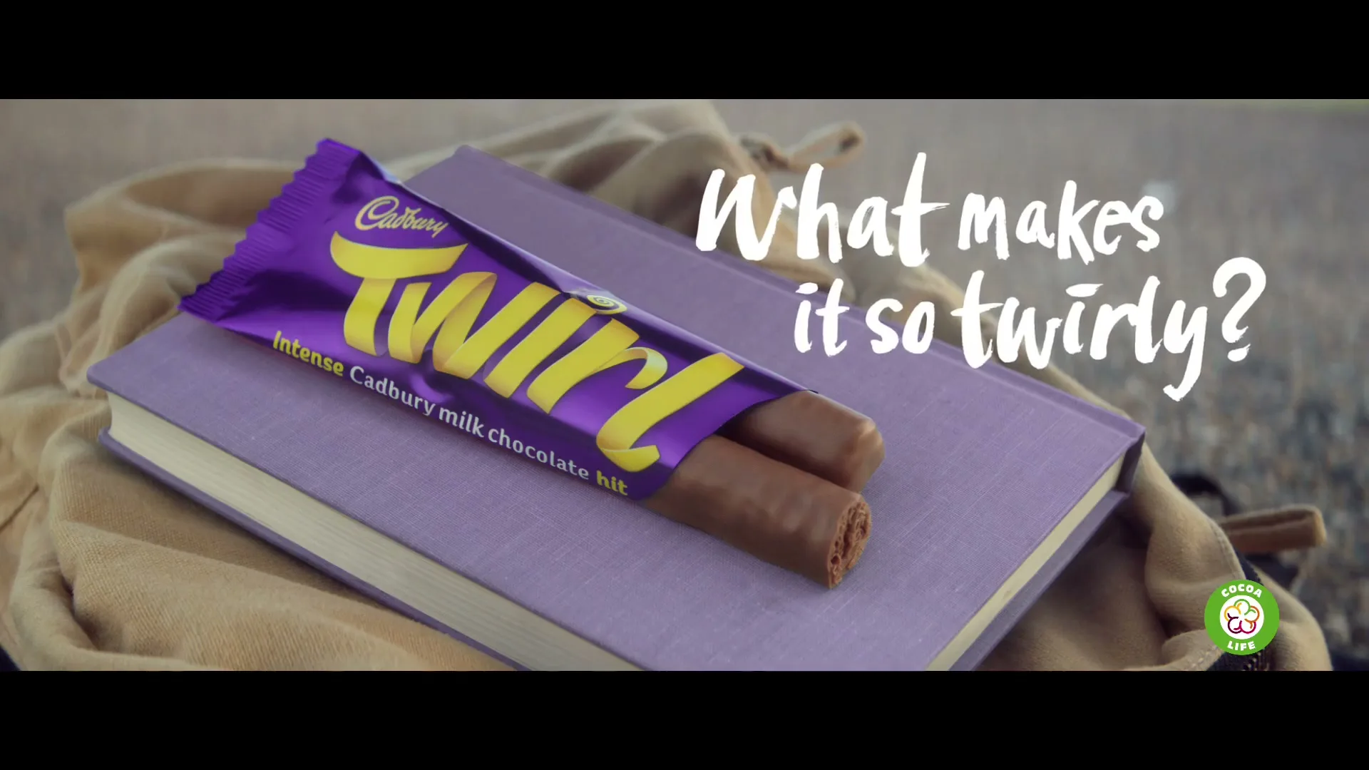 A Cadbury Twirl chocolate bar, unwrapped, rests on a purple book, with a beige backpack in the background, asking "What makes it so twirly?"