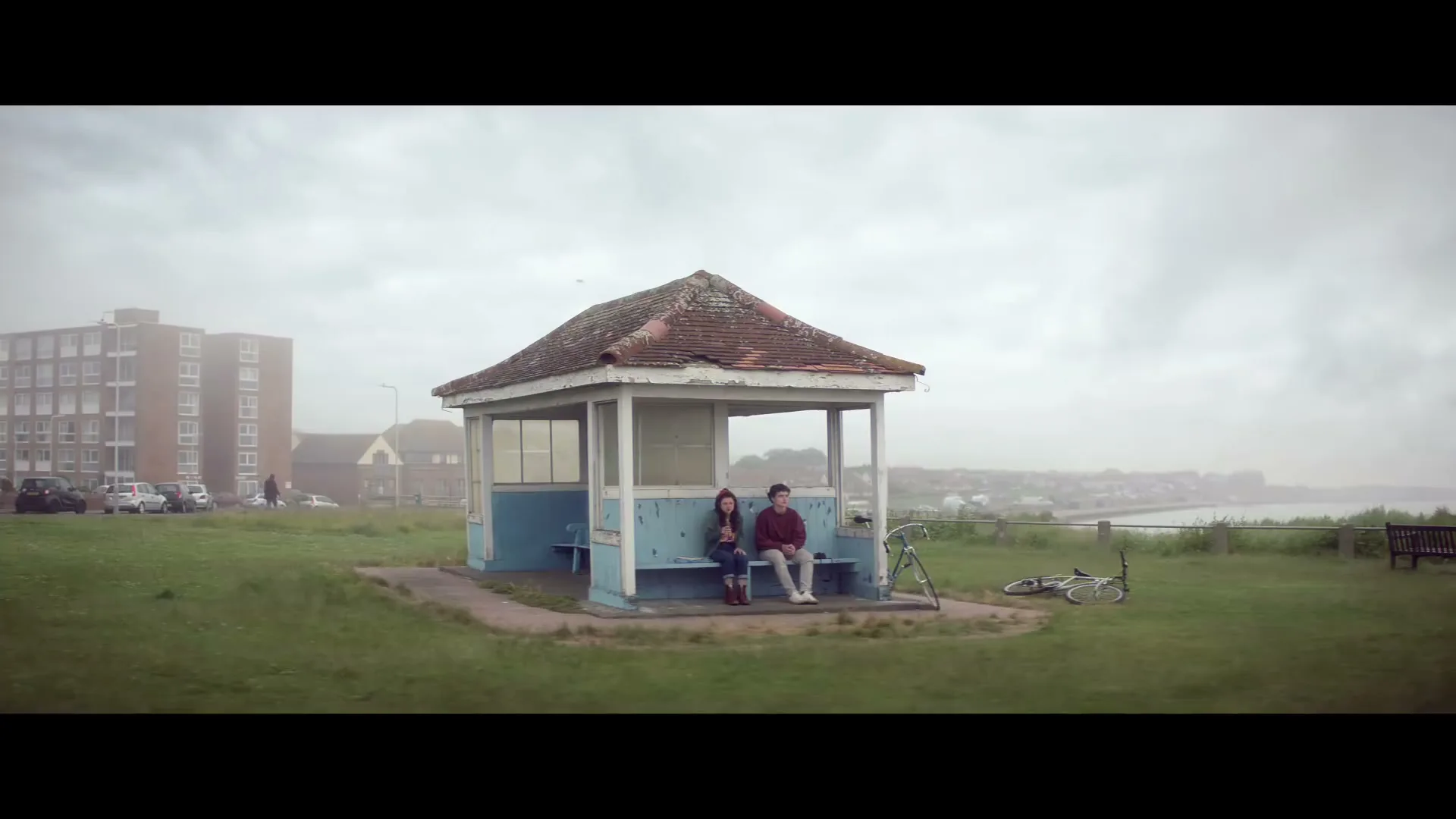A young couple sits on a bench inside a small, weathered blue gazebo on a grassy hill overlooking the ocean on a cloudy day.