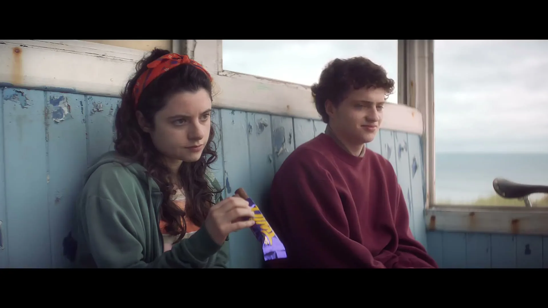 A young woman with a red headband and a young man sit on a weathered blue bench, looking out at the ocean on a cloudy day.