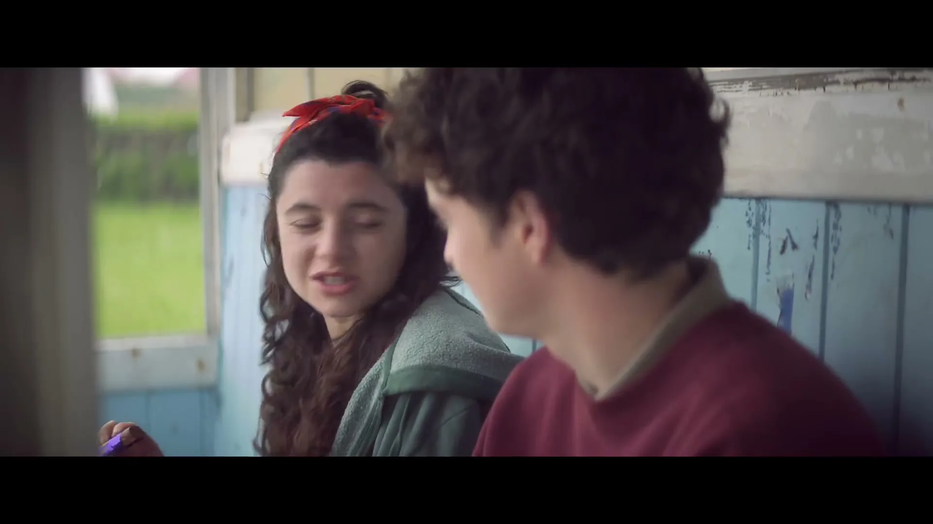 A young woman with dark curly hair and a red headband talks to a young man in a maroon shirt, sitting in a rustic, light blue wooden interior.
