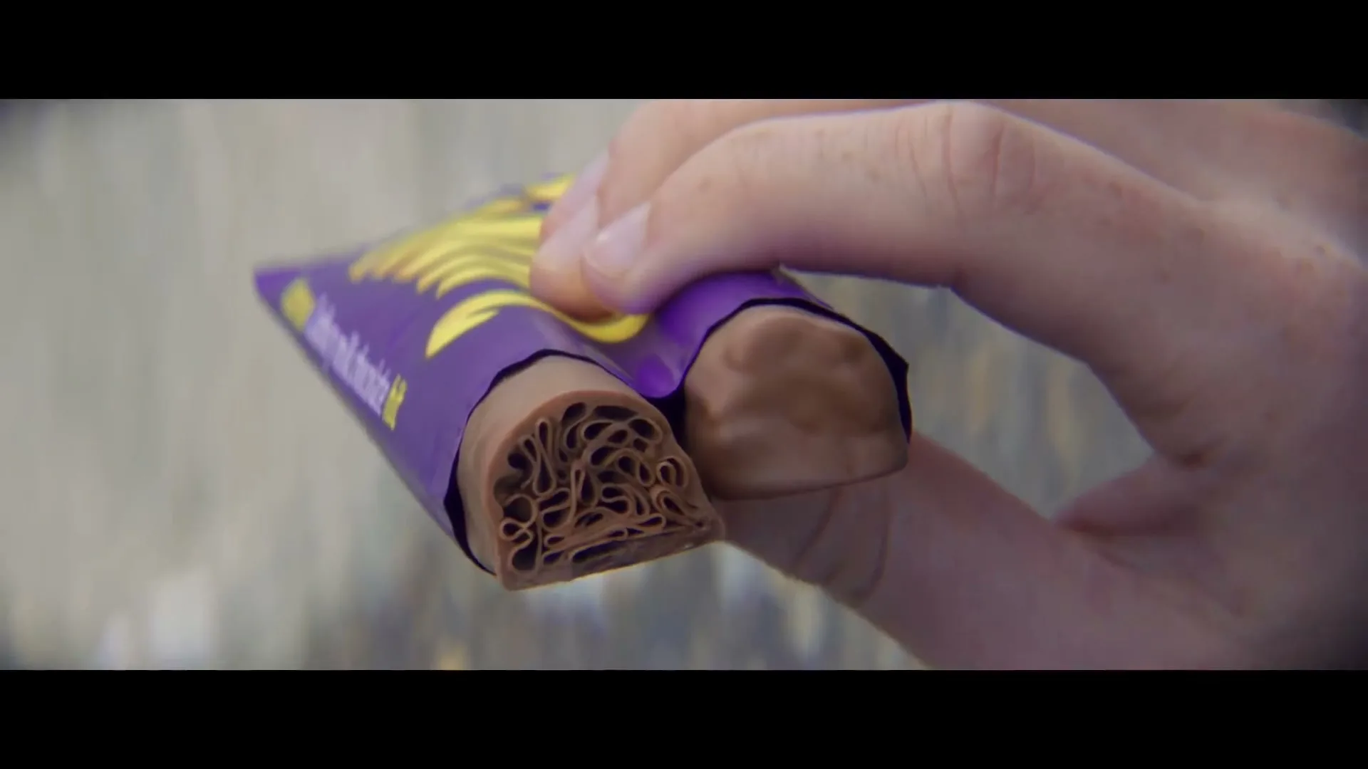 A hand holds a Cadbury chocolate bar, revealing its bubbly interior, against a blurred background.