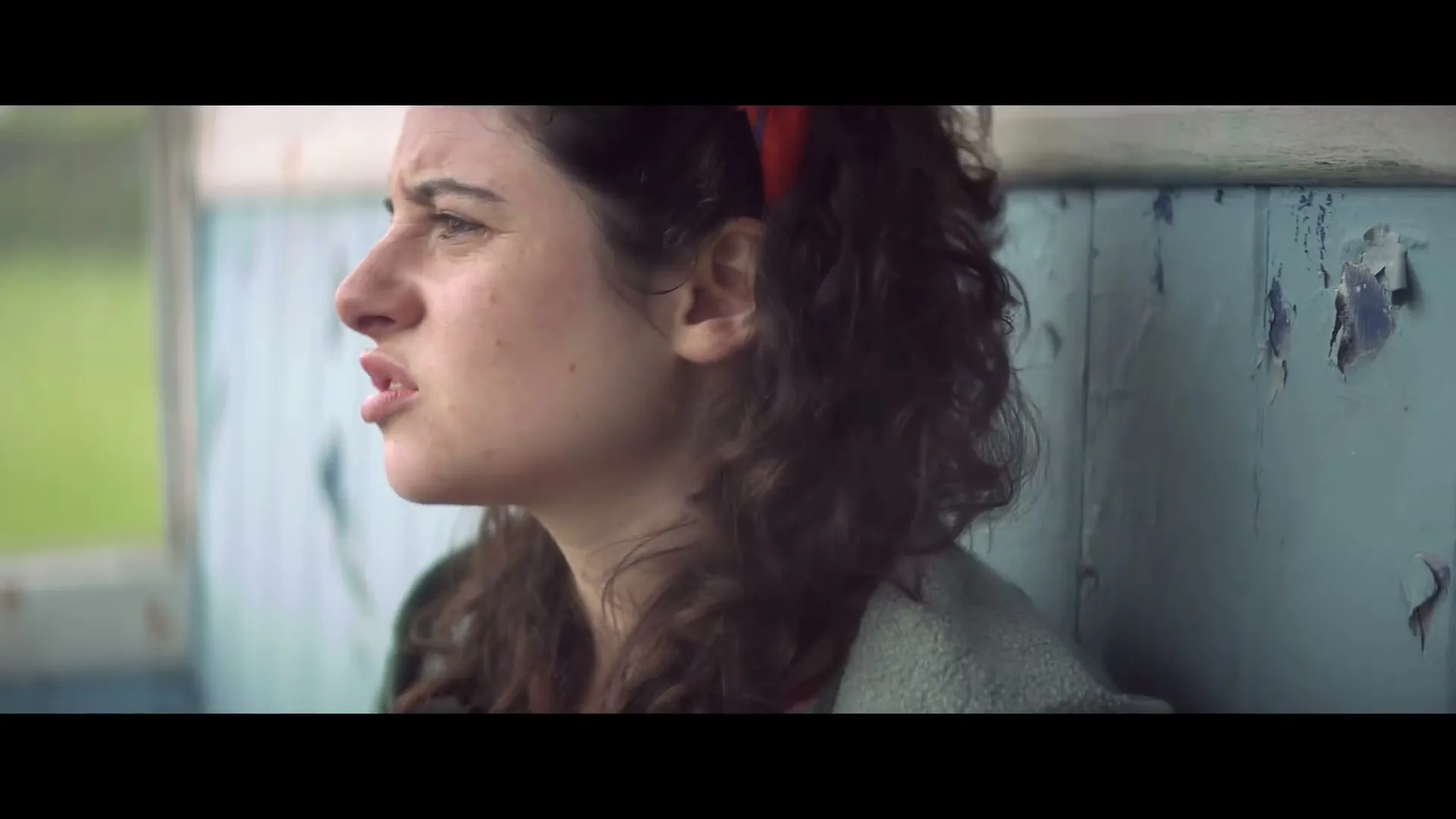 A young woman with curly hair and a red headband looks off-screen, speaking, while sitting against a peeling blue wall.
