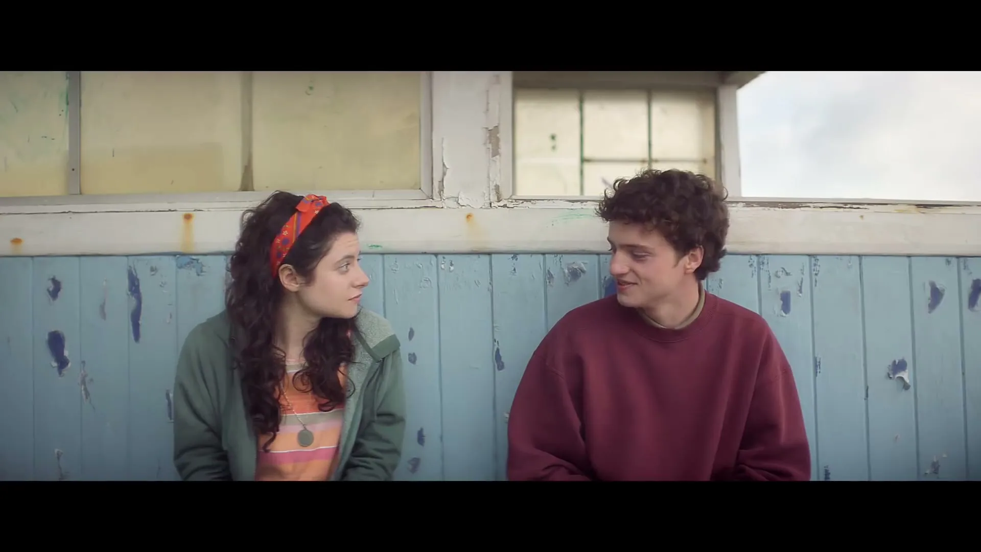A young woman with curly hair and a young man with curly hair sit side-by-side on a weathered blue bench, looking at each other and talking.
