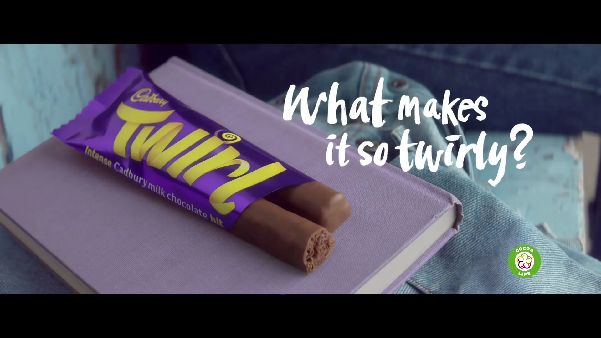 A Cadbury Twirl chocolate bar, partially unwrapped, rests on a light purple book, with a person's denim-clad leg visible in the background.