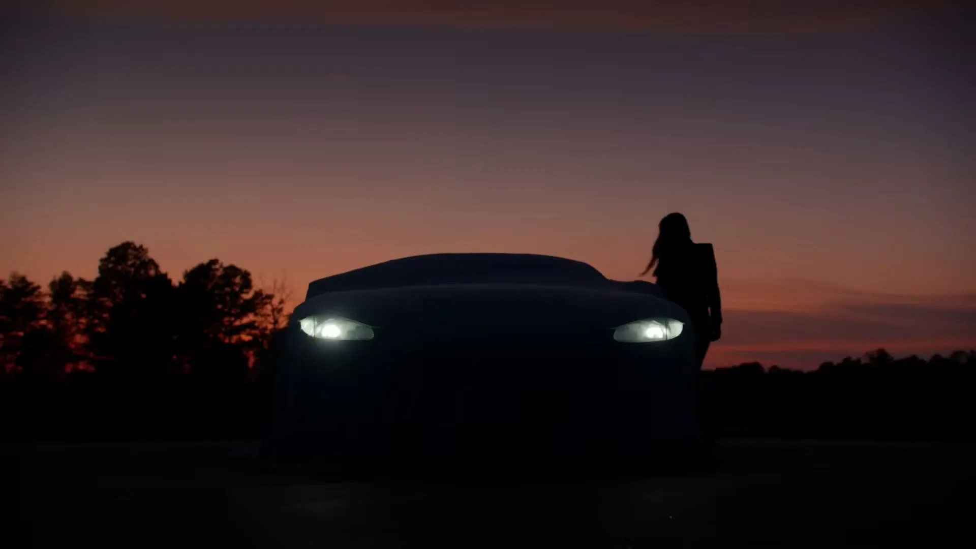 A woman in silhouette stands next to a car with its headlights on, against a dramatic dusk sky with trees in the background.