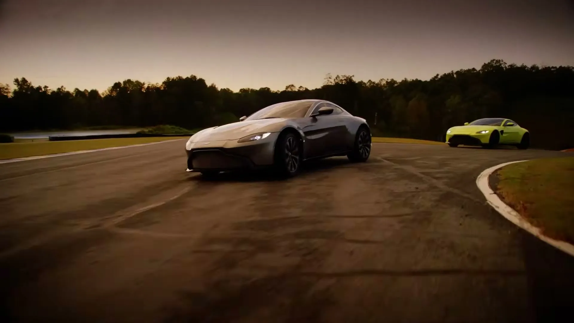 Two Aston Martin sports cars, one grey and one lime green, drive on a race track at dusk, with trees in the background.