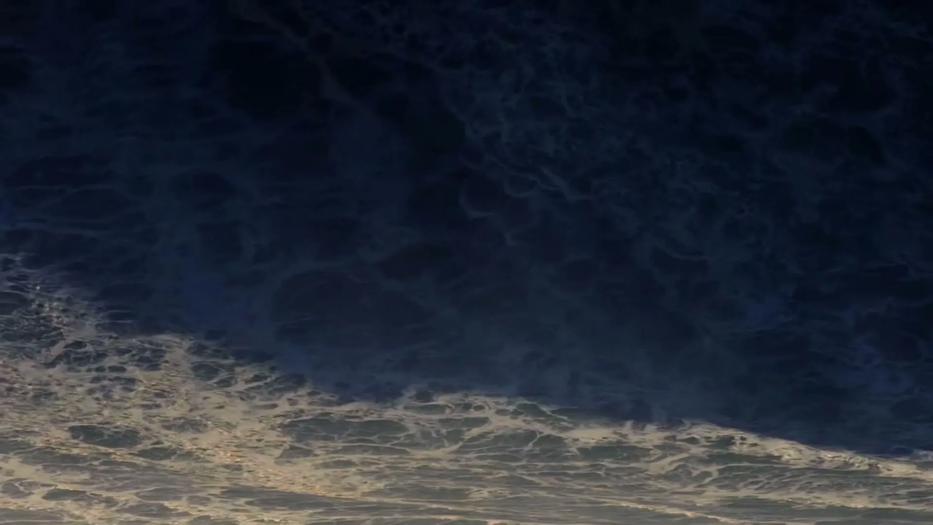 An aerial shot captures the dark blue ocean with white foamy waves crashing, creating a dynamic and powerful natural scene.