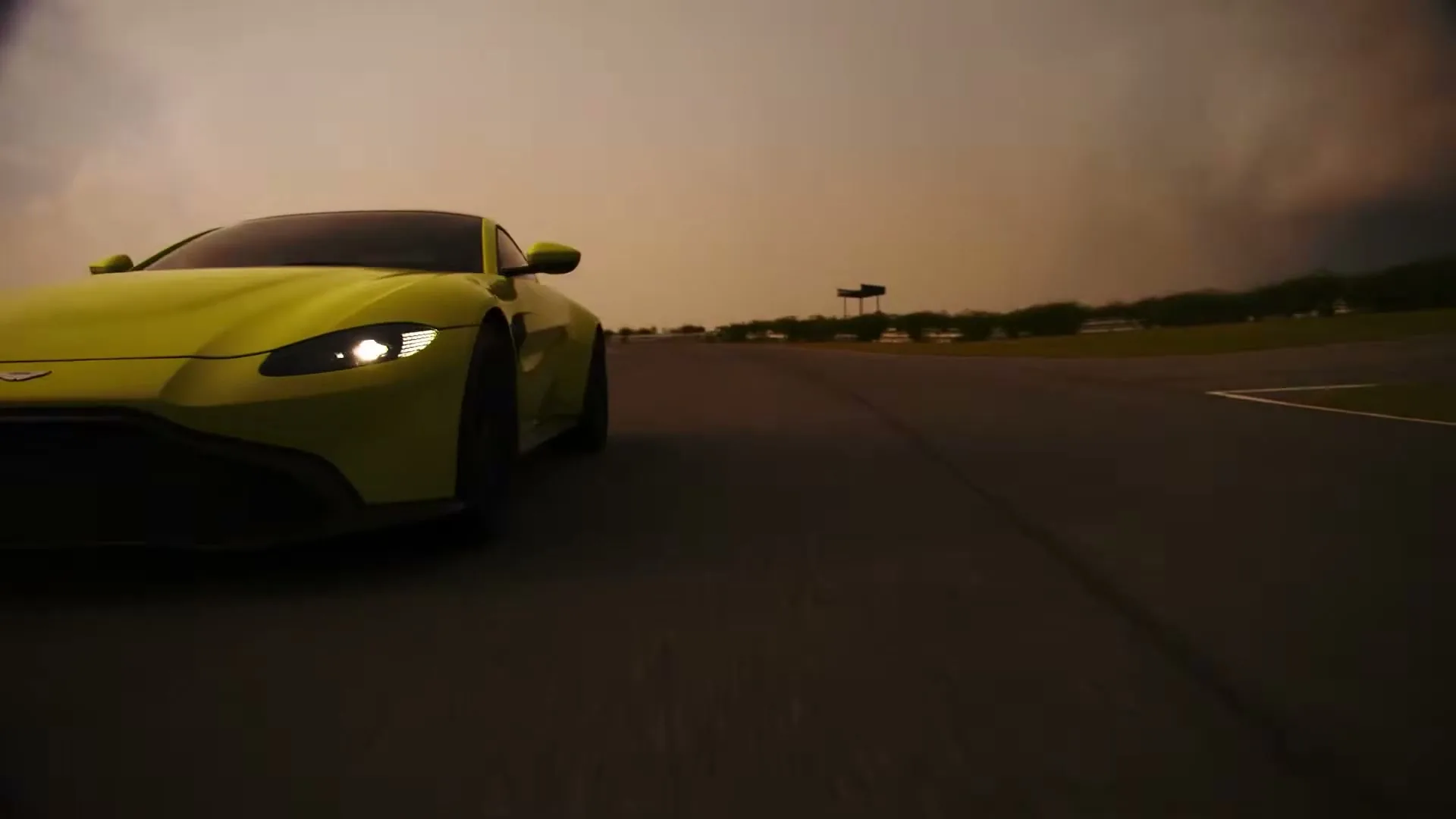 A bright yellow Aston Martin Vantage drives quickly on a race track under a dramatic, cloudy sky at dusk.