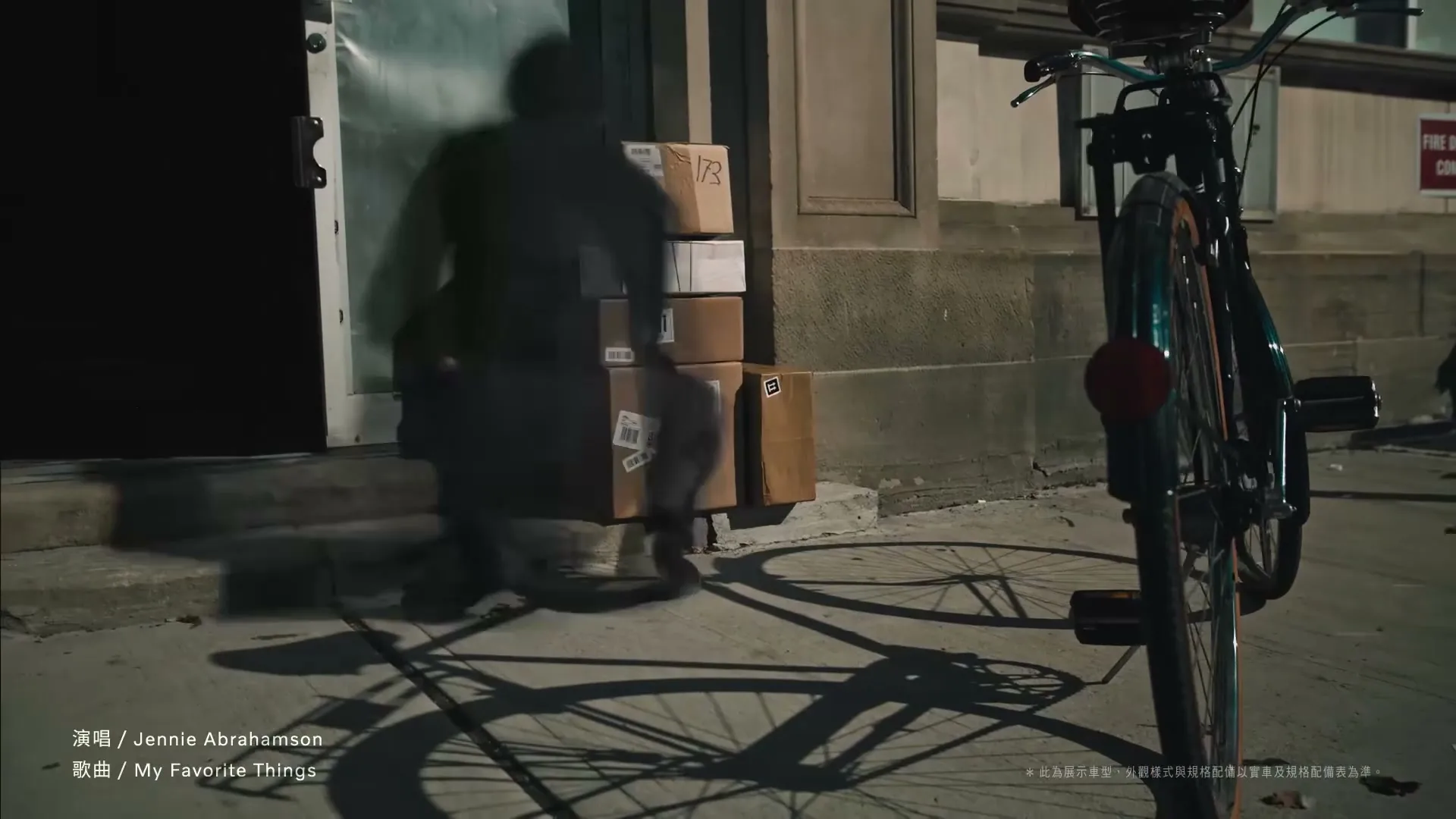 A person quickly stacks many cardboard boxes on a city sidewalk next to a parked bicycle, with sunlight casting long shadows.