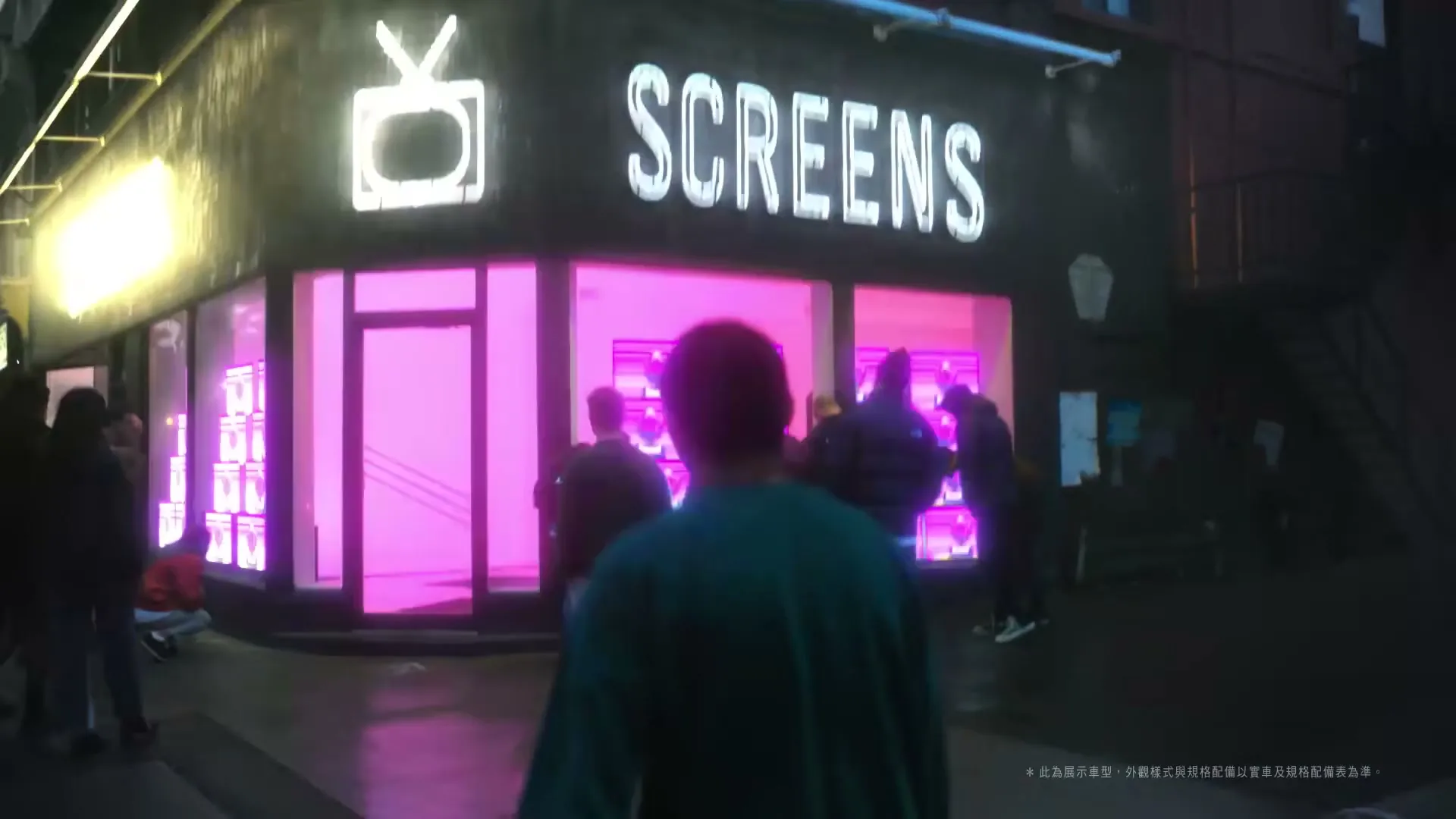 A person in a green sweatshirt walks away from the camera towards a store with "SCREENS" in neon lights, as other people mill about on the street at night.