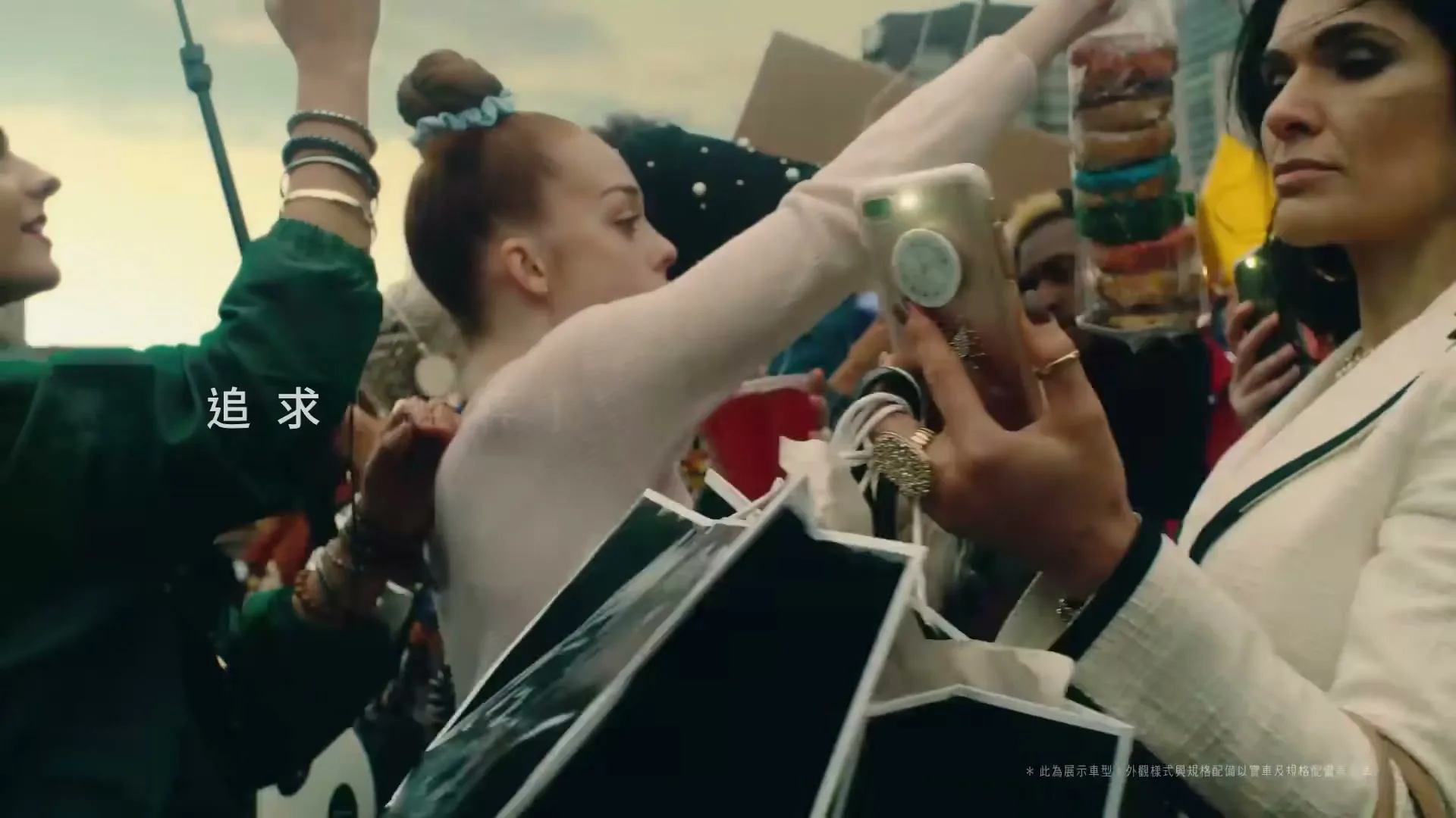 A diverse group of people are seen in a crowd, with a woman in a white jacket holding up a phone to take a picture, while another woman in a pink top holds up a bag of colorful donuts.