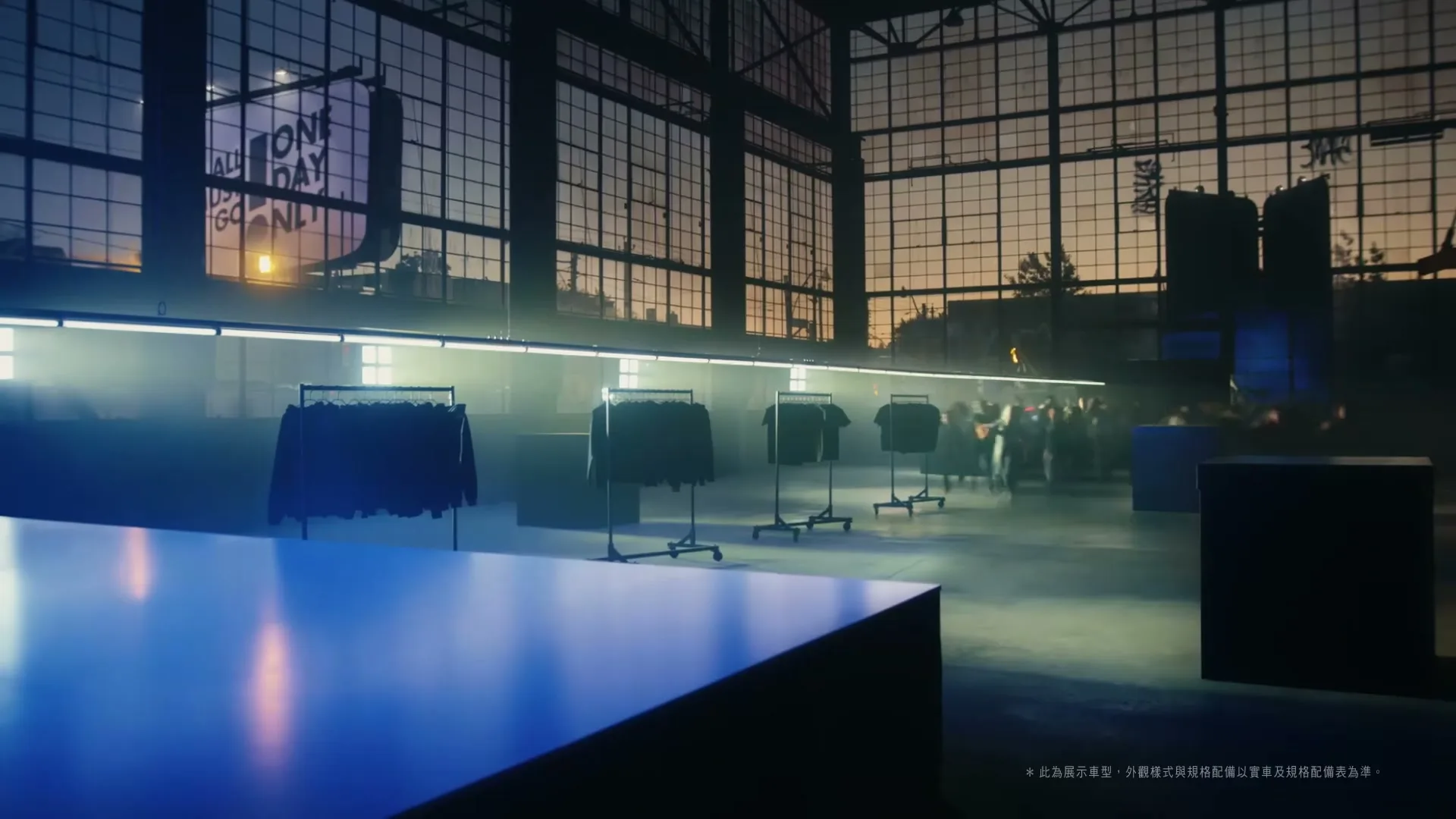 A wide shot of a large, empty industrial space with clothes racks and a long table, as a crowd of people rushes in from the right.
