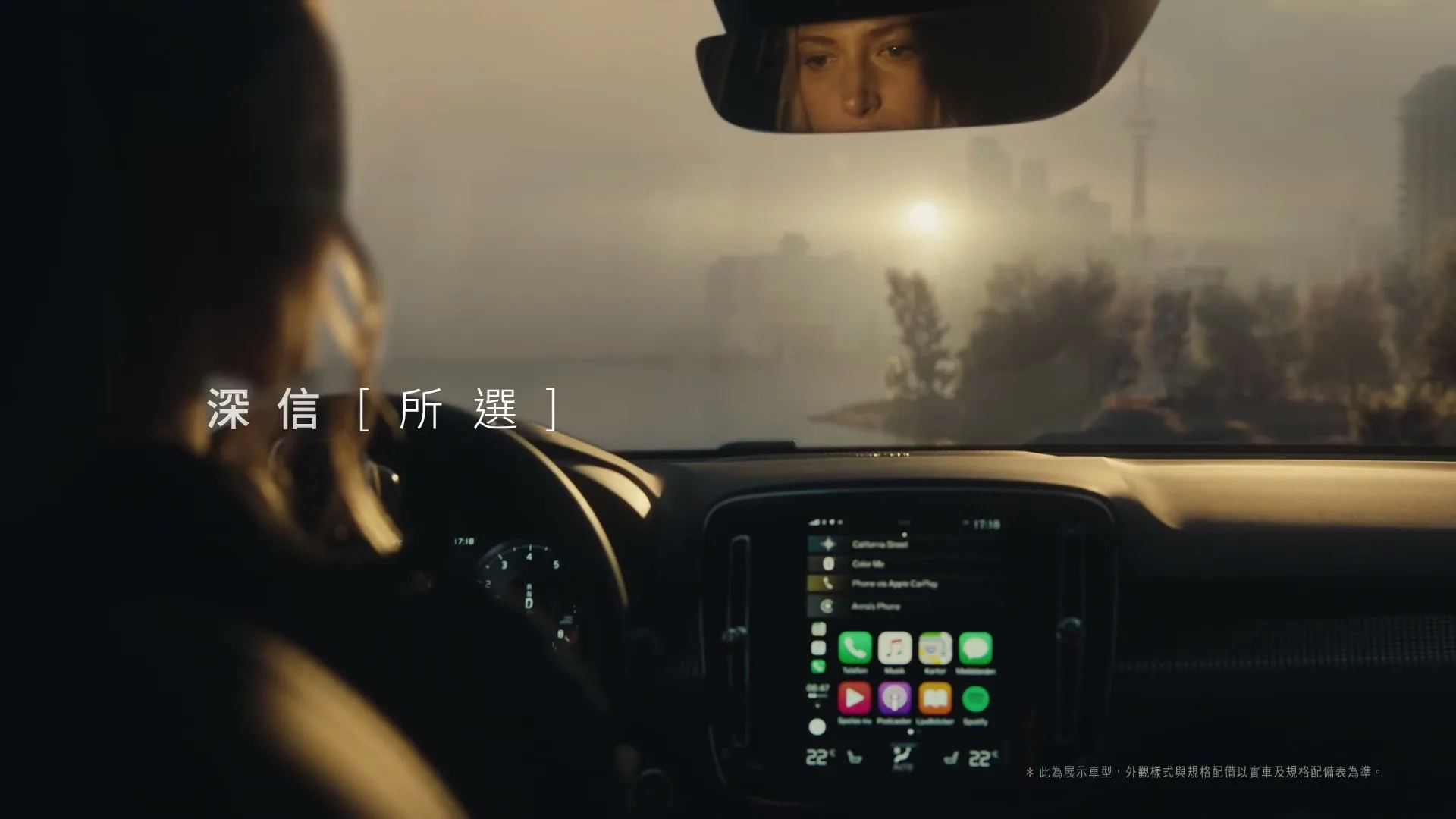 A woman drives a Volvo car at golden hour, with her reflection visible in the rearview mirror and a city skyline in the background.