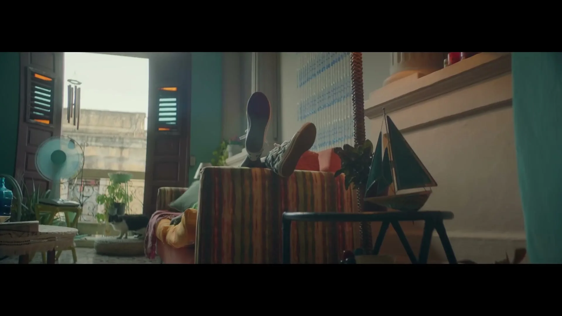 A person in a green shirt and sneakers is seen relaxing on a striped couch with their feet up, in a colorful living room with an open door leading to a balcony.