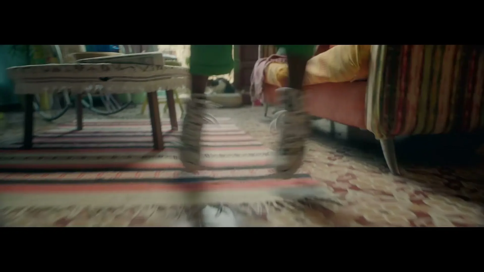 A person wearing green pants and patterned sneakers jumps up and down on a patterned rug in a colorful, bohemian-style living room.