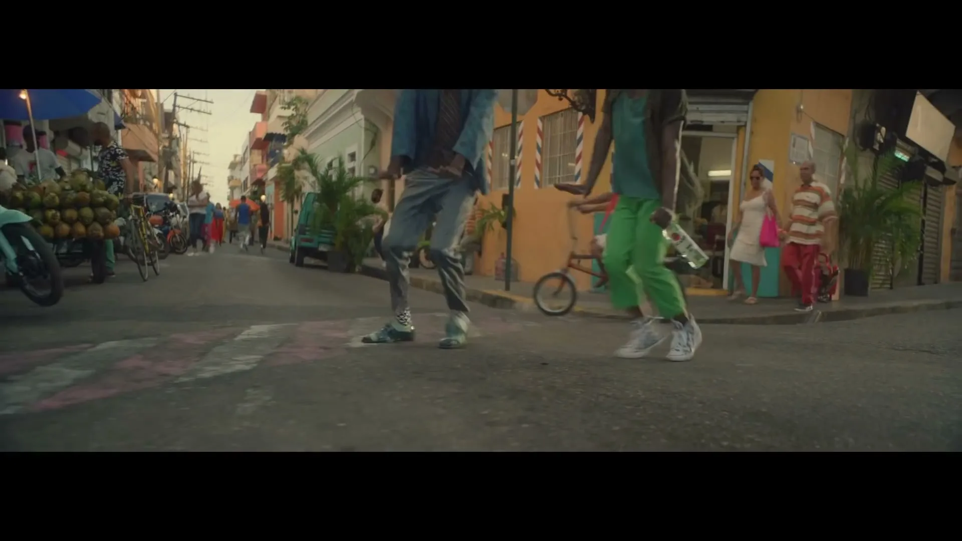 Two men in casual clothes dance in the street, with other people and buildings in the background, during golden hour.