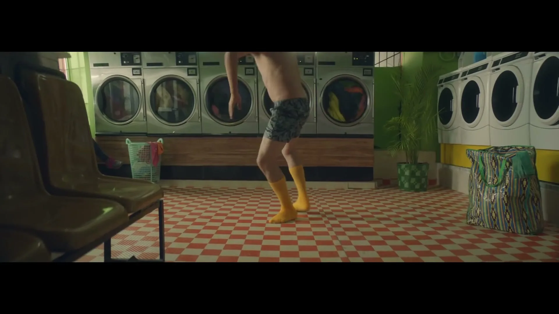 A shirtless man in patterned shorts and yellow socks dances in a laundromat with washing machines and yellow chairs.