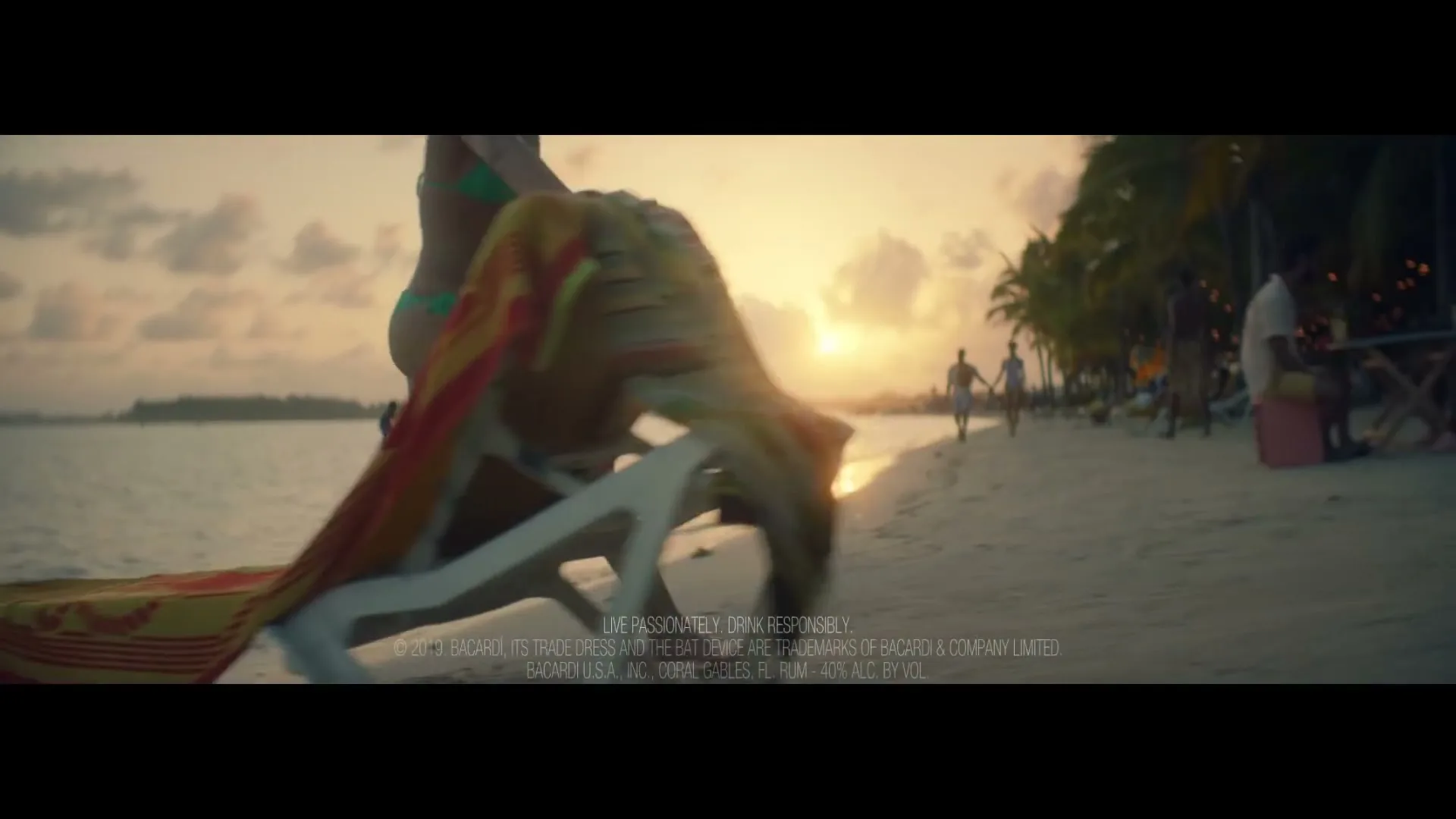A woman in a green bikini walks past a beach chair on a sandy beach at sunset, with other people in the background.