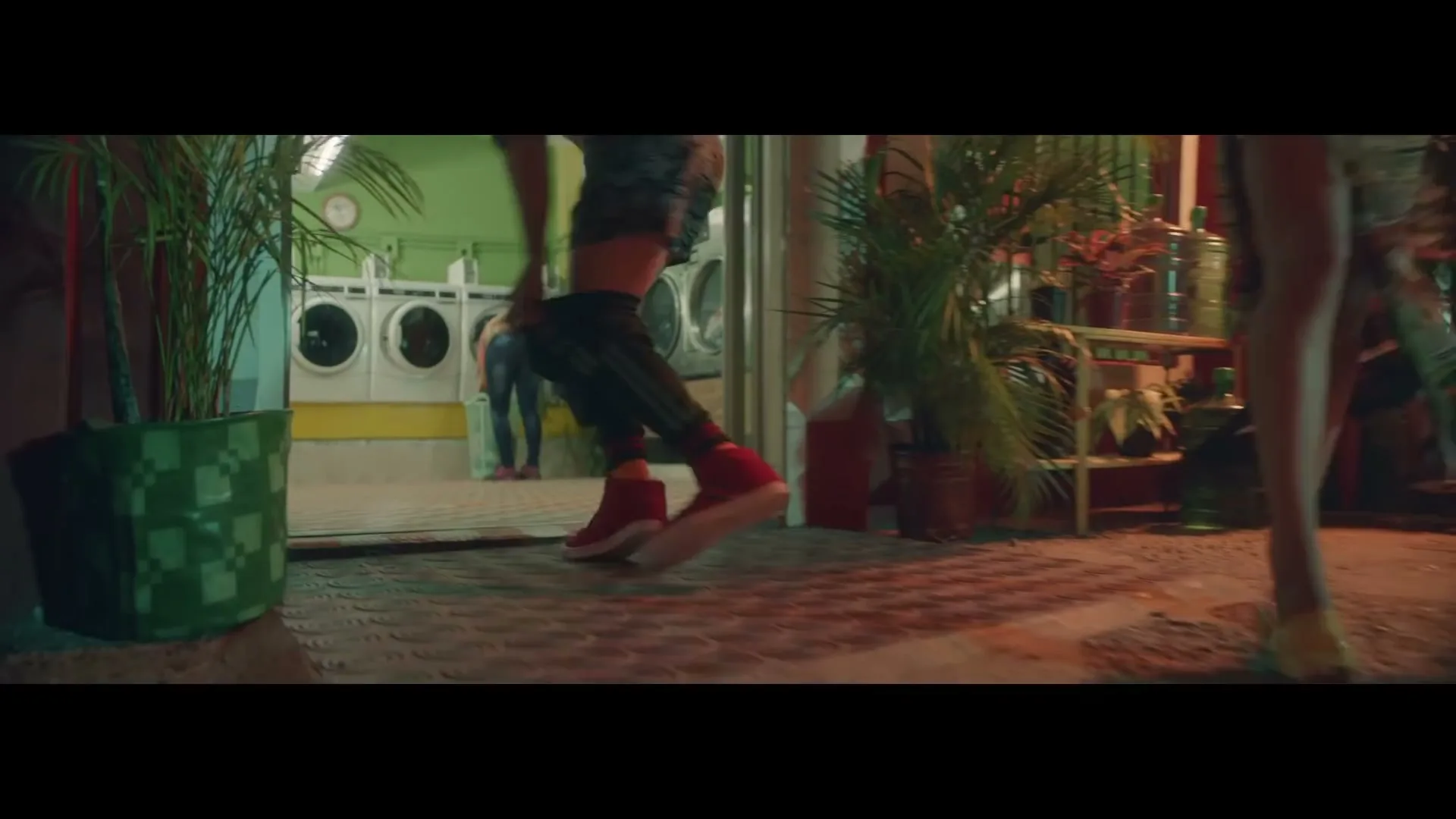 A person in red shoes dances in front of a laundromat at night, with other people visible in the background.