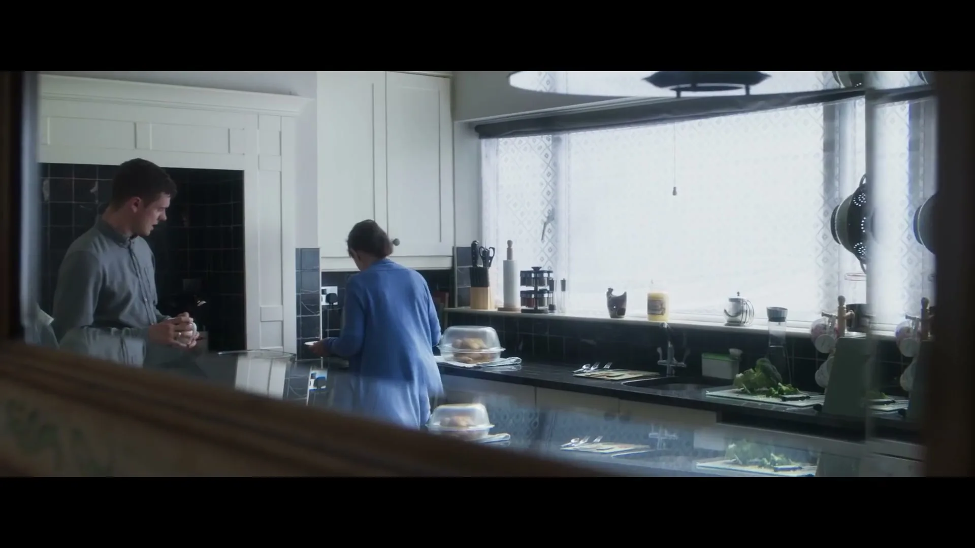 A young man and an older woman are in a bright kitchen, with the woman preparing food on the counter while the man holds a mug.