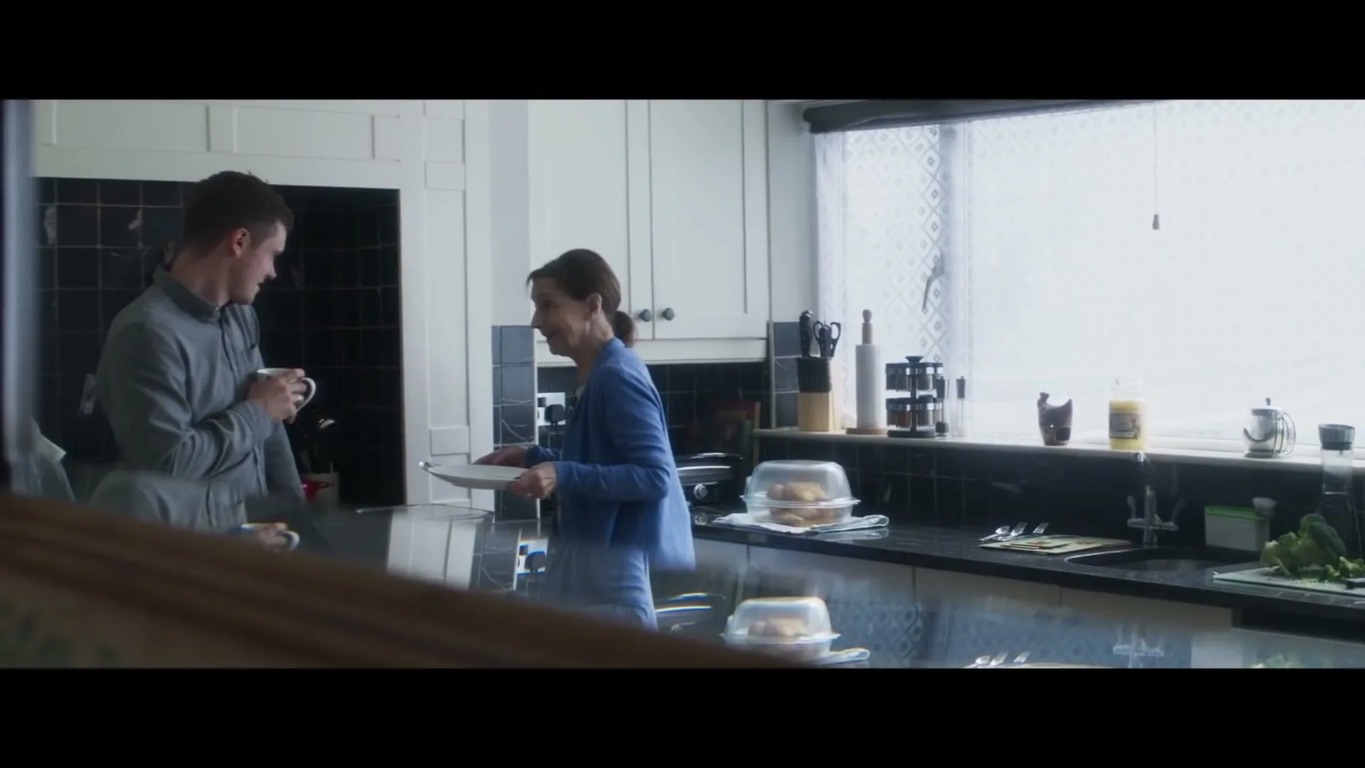 A young man in a grey shirt holds a mug and talks to a woman in a blue cardigan who is setting a plate on a kitchen counter.