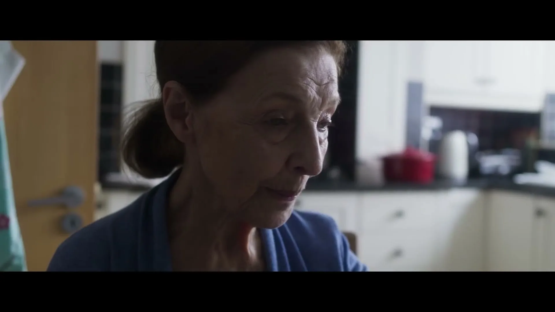An older woman with brown hair in a blue top looks down and smiles slightly in a kitchen.