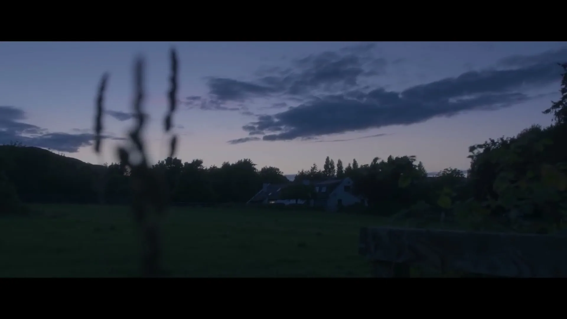 A wide shot of a rural landscape at dusk, with a house nestled among trees in the distance and blurred foliage in the foreground.