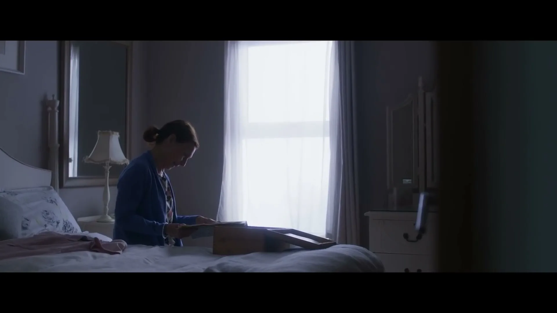 A woman in her 40s sits on a bed, looking at old photographs in a box, in a dimly lit bedroom.