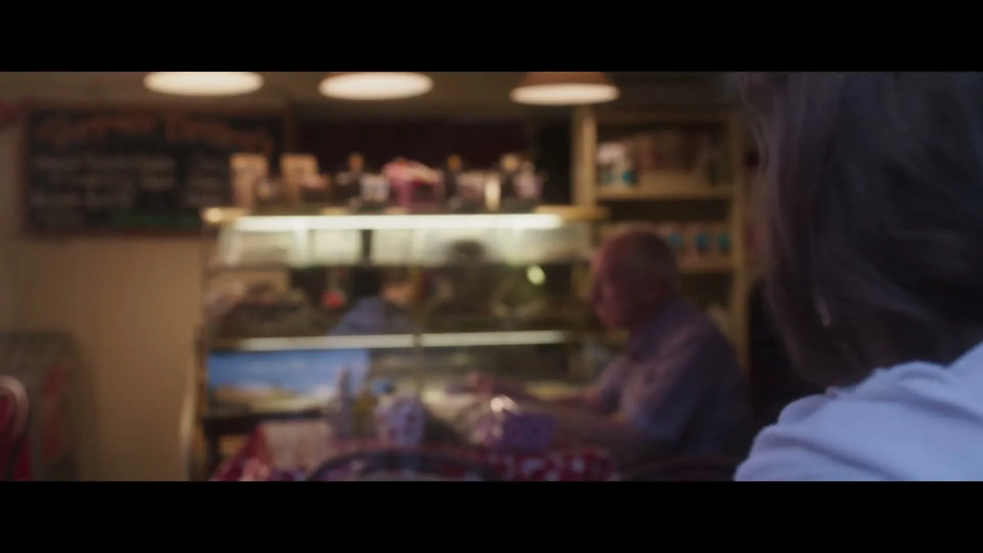 An elderly man in a plaid shirt sits at a table in a cafe, looking at a bouquet of flowers, with a dessert display case behind him.