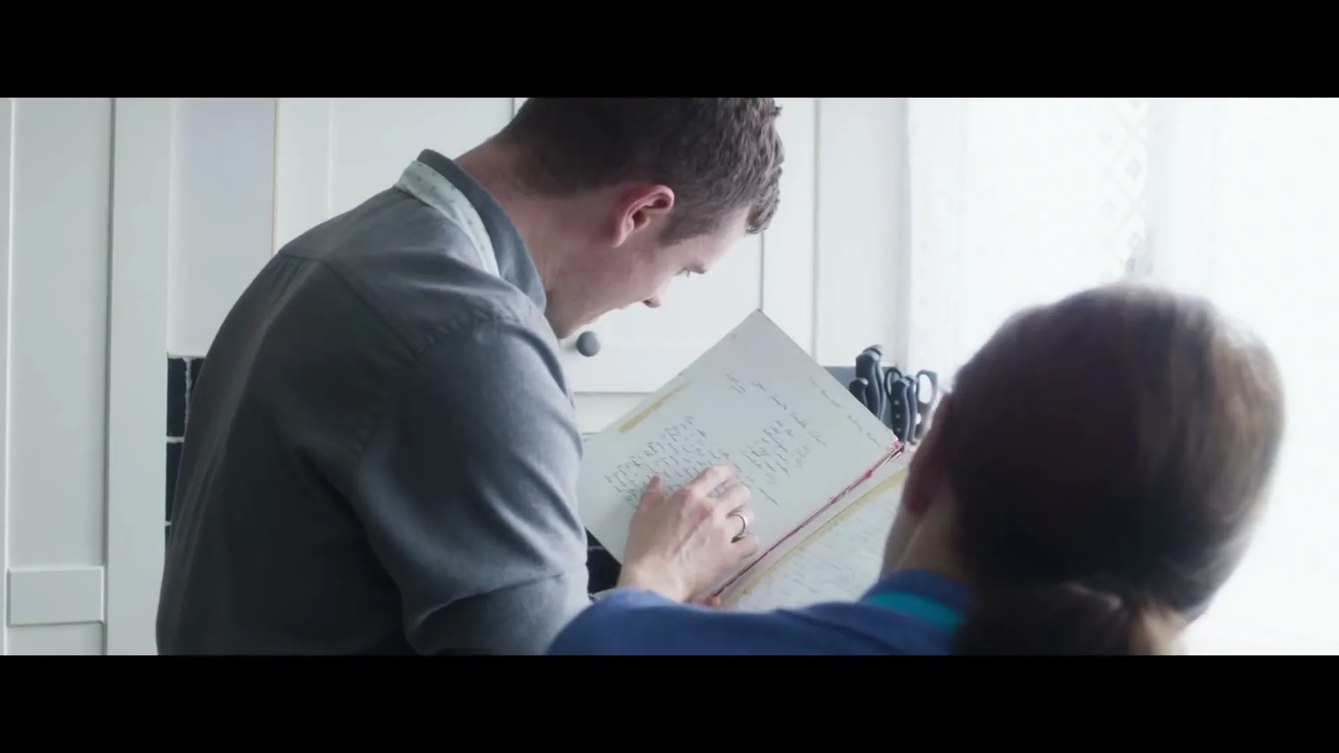 A man and a woman stand in a bright kitchen, looking at a handwritten recipe book together, with the man pointing at the page.