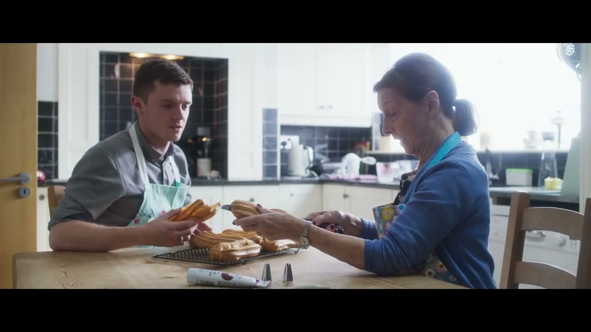 A young man and an older woman, both wearing aprons, sit at a wooden table in a bright kitchen, decorating eclairs.