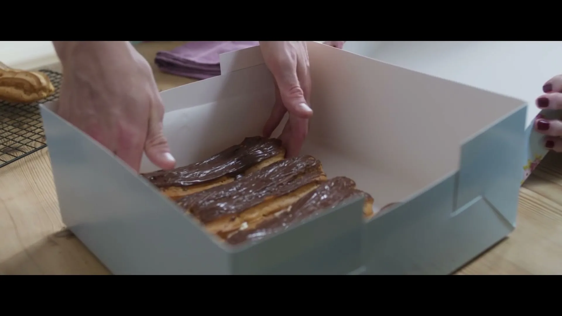 A person's hands carefully place chocolate eclairs into a light blue and white box on a wooden table.