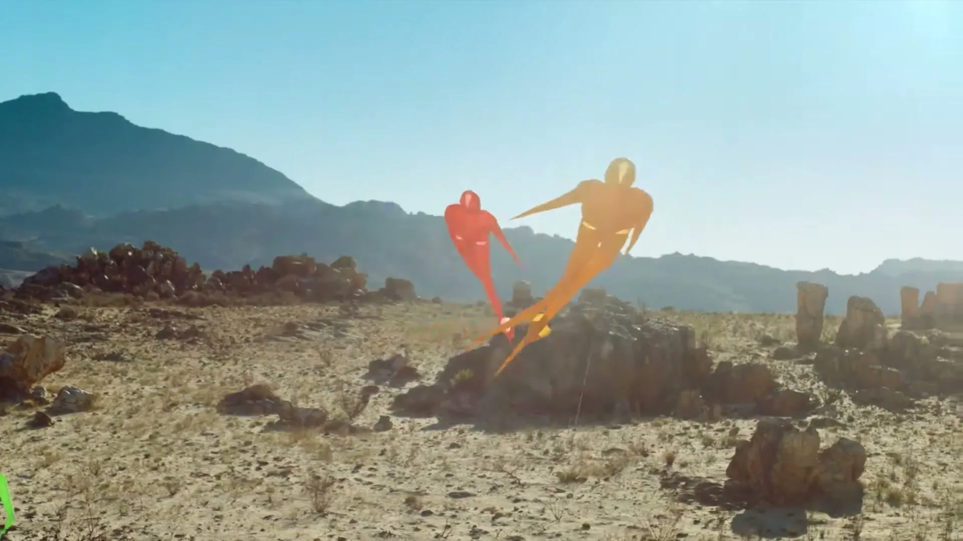 Two abstract, human-shaped kites, one orange and one red, fly over a rocky, arid landscape under a clear blue sky.