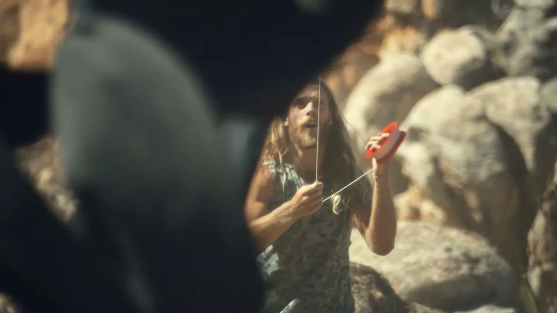 A man with long hair and a beard, wearing a tank top, plays with a yo-yo in a rocky, outdoor setting.