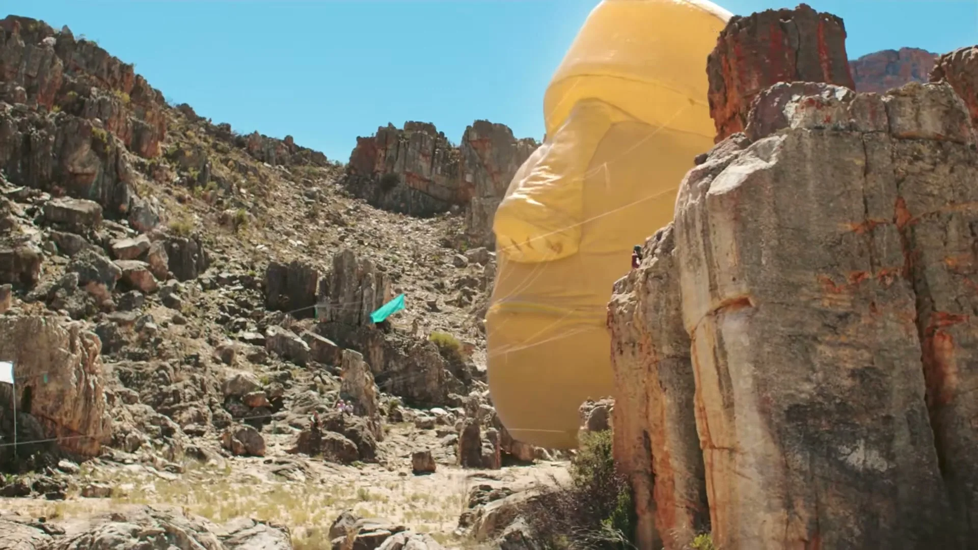 A giant inflatable head, resembling a Moai statue, is being pulled through a rocky, arid canyon under a clear blue sky.
