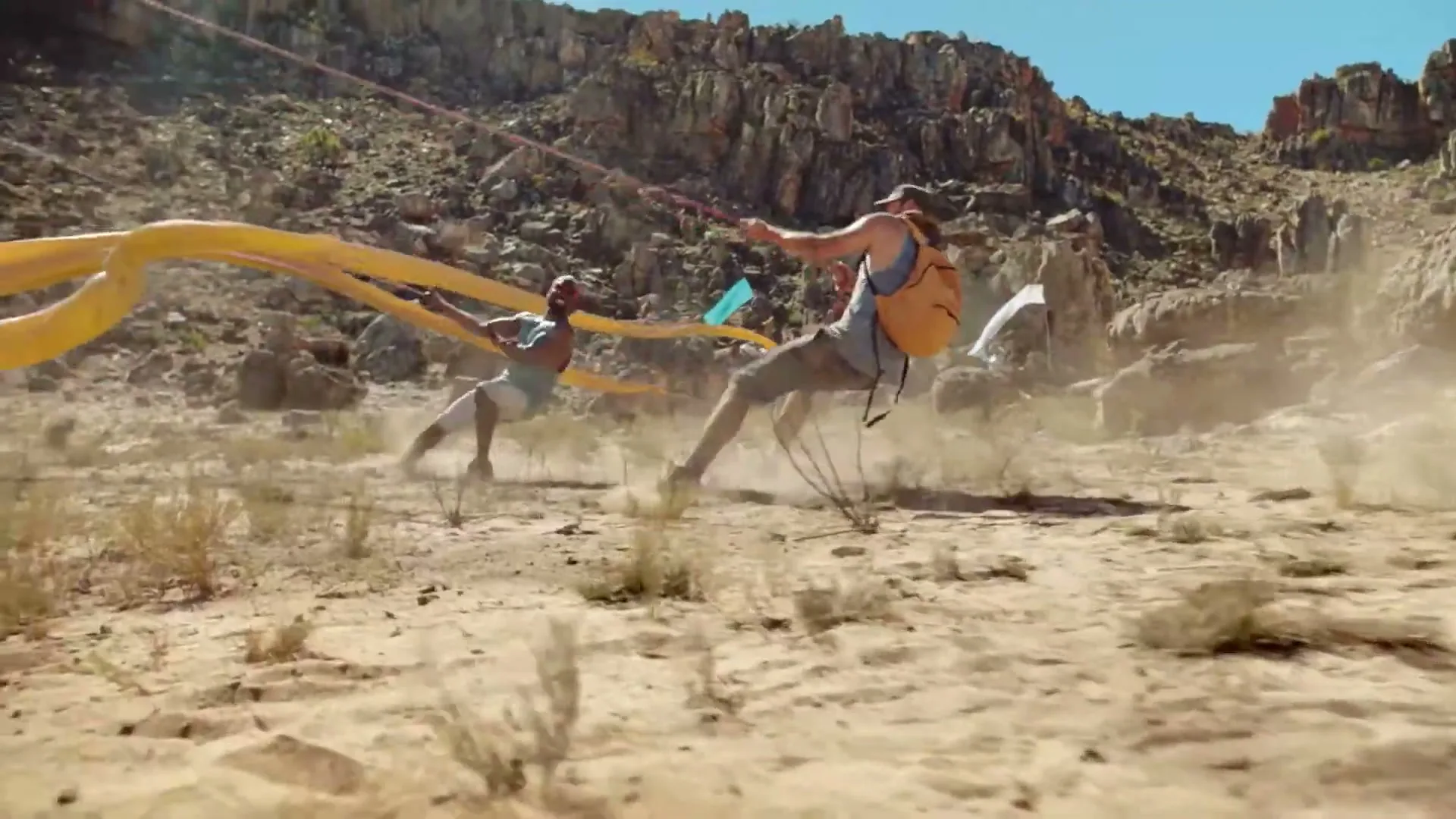 Two men in casual clothes are pulled by ropes, kicking up dust in a rocky, arid landscape under a bright sky.