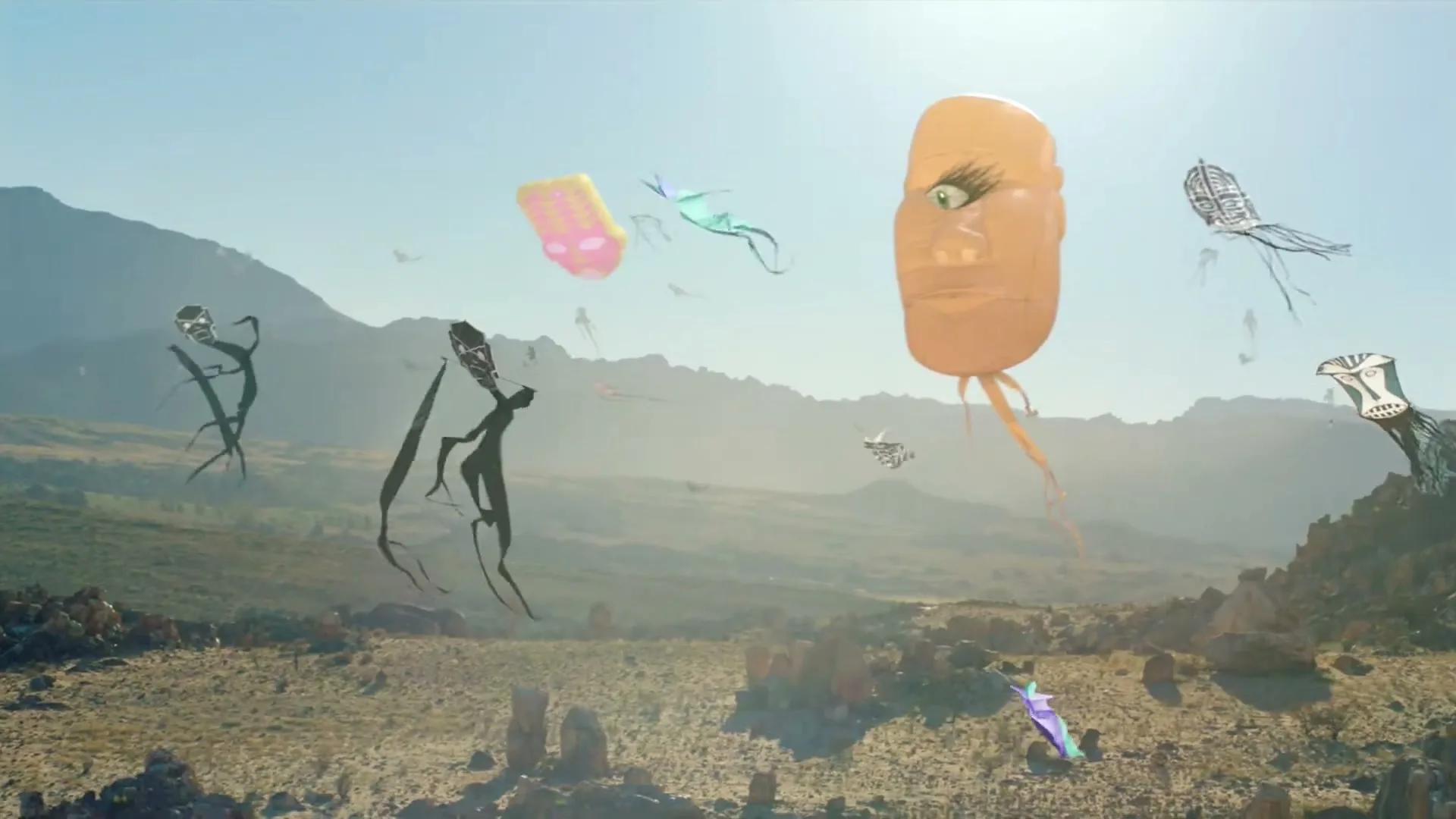A wide shot of a desert landscape with mountains in the background, as various colorful, abstract kites fly in the sky under a bright sun.