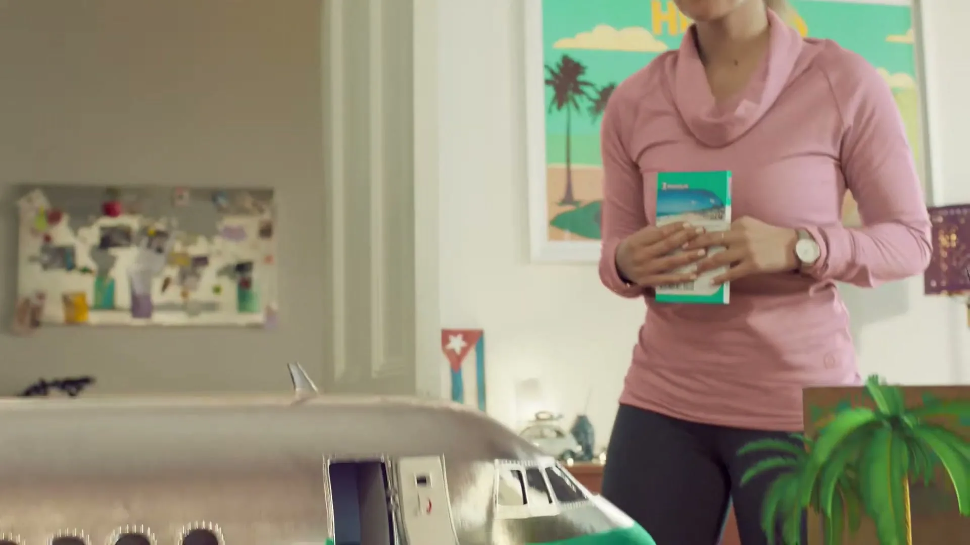 A young woman in a pink top holds a brochure, as a miniature version of her exits a toy plane and gets into a toy car.