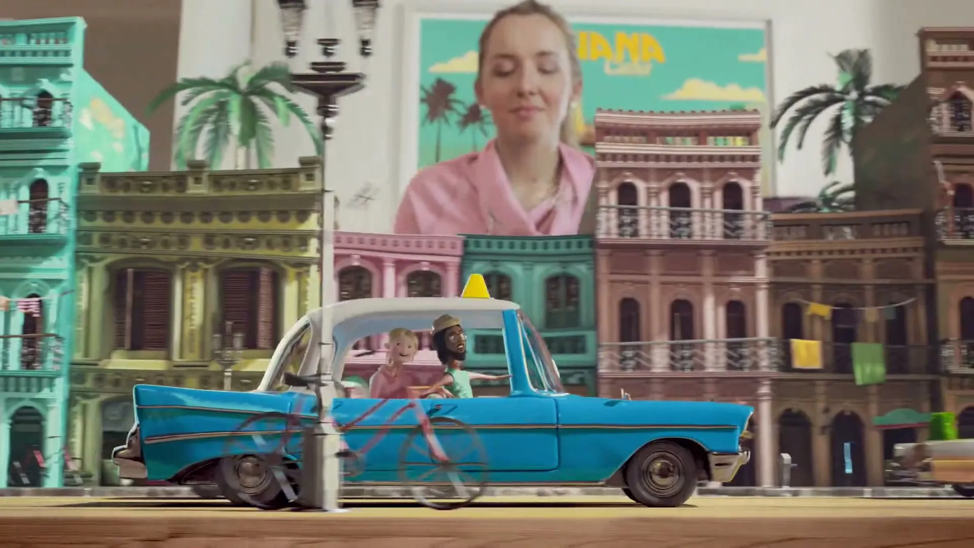 A woman in pink pajamas smiles behind a miniature diorama of Havana, Cuba, with a blue toy car driving past colorful buildings.