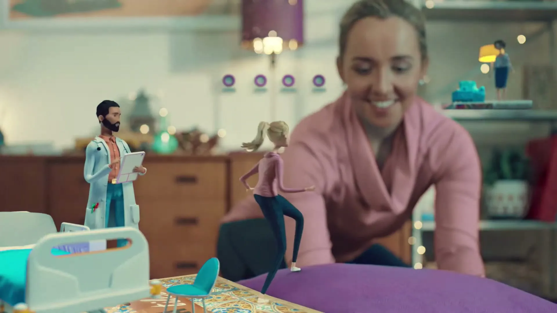 A smiling woman in a pink top is seen from the chest up, looking down at a miniature hospital scene with a doctor and patient doll.