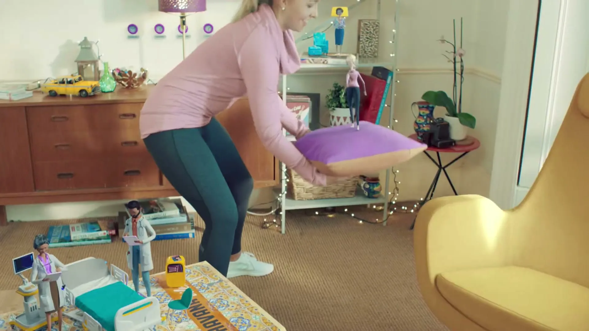 A woman in a pink top and dark leggings carefully places a purple cushion with a small doll on it, next to miniature hospital figures on a rug in a living room.