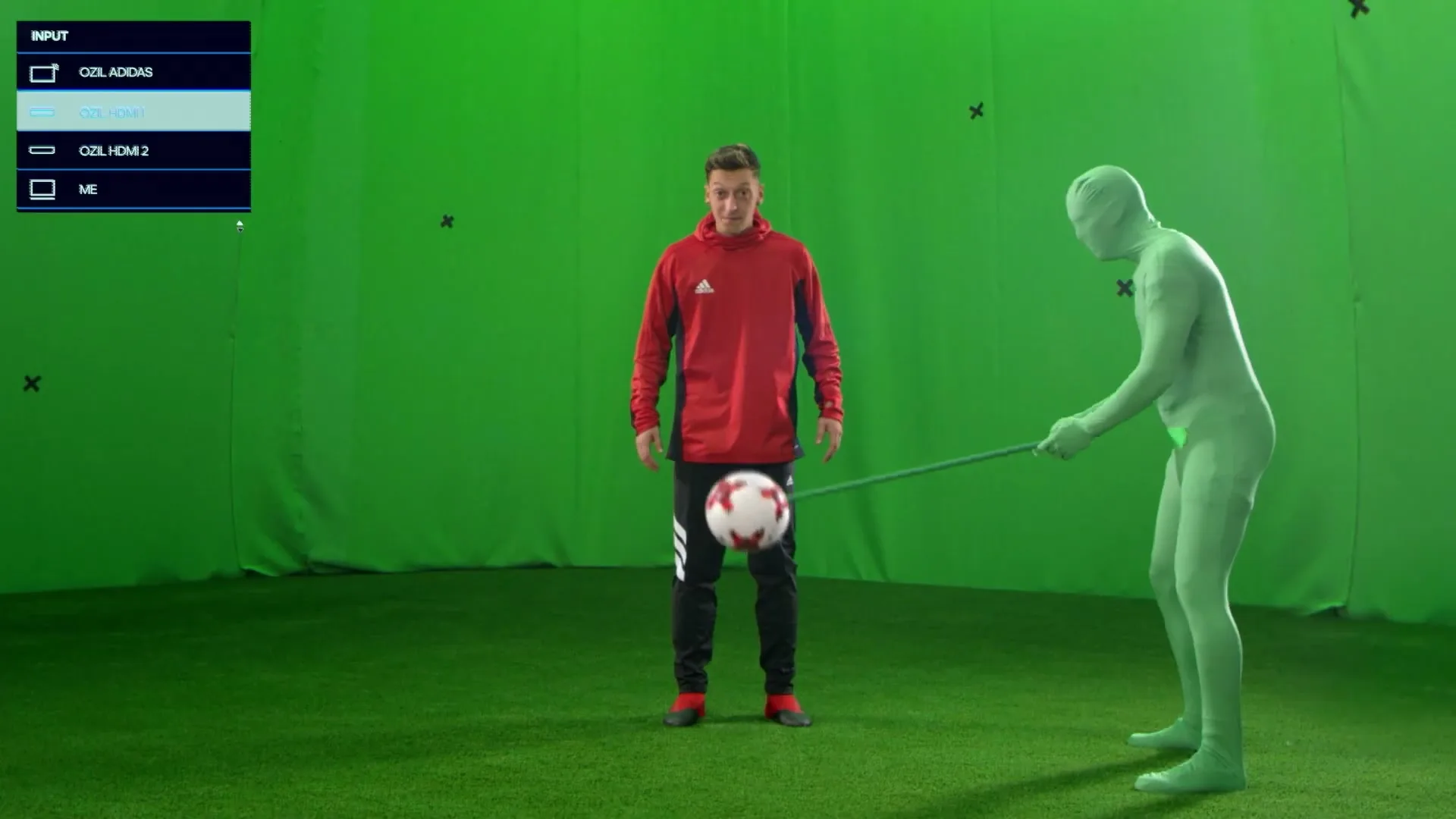 A male soccer player in a red jacket juggles a soccer ball while a person in a green screen suit holds a stick near the ball, all on a green screen set.