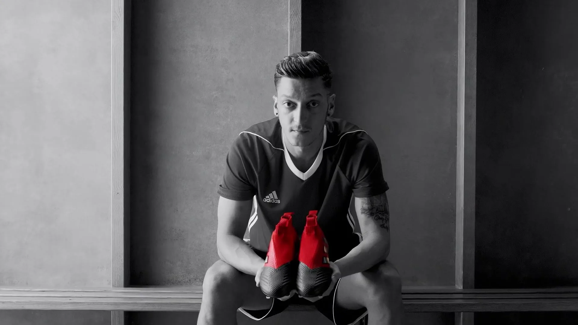 Mesut 
Ozil, a male soccer player, sits on a bench in a locker room, holding a pair of red soccer cleats.
