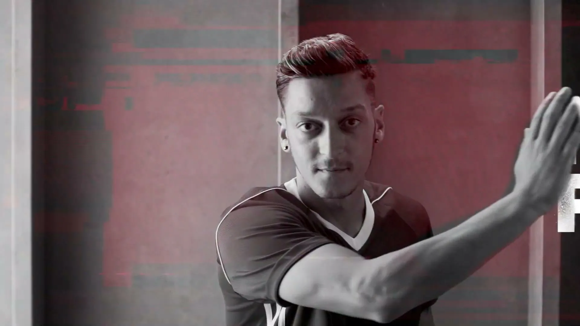 Mesut Özil, a male soccer player, in a black and white shot with red accents, waves his hand in front of a red wall with text.