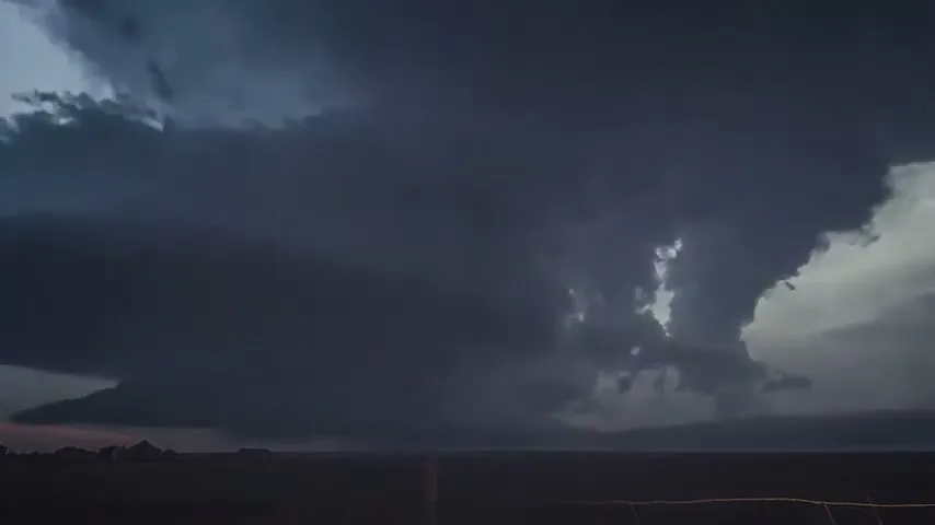 A wide shot of a dramatic, dark storm cloud formation with multiple lightning strikes illuminating the sky over a flat, open landscape at dusk.