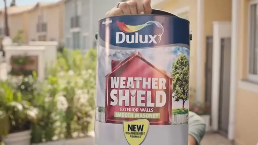 A woman with dark hair smiles at the camera, then a can of Dulux Weather Shield paint is raised in front of her face in a suburban setting.