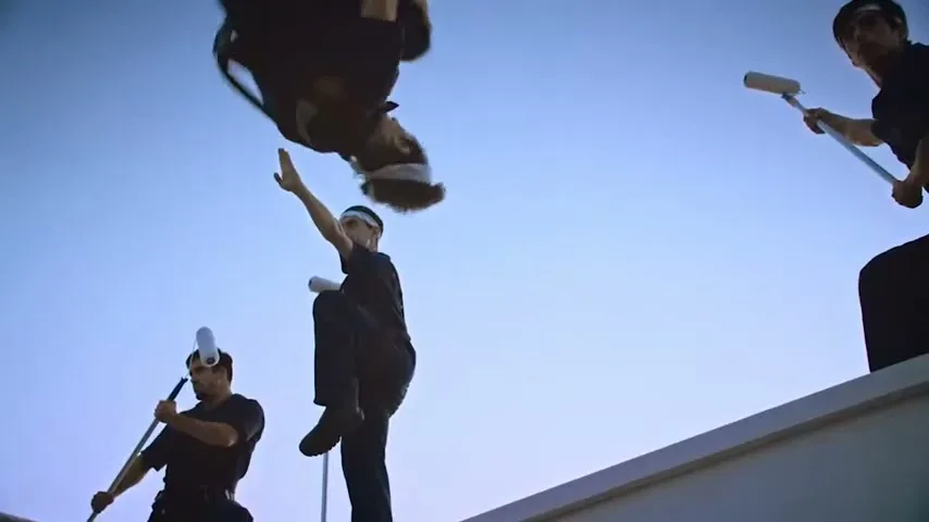 Four male painters in black uniforms and white headbands are on a white rooftop, with one performing a backflip against a clear blue sky.