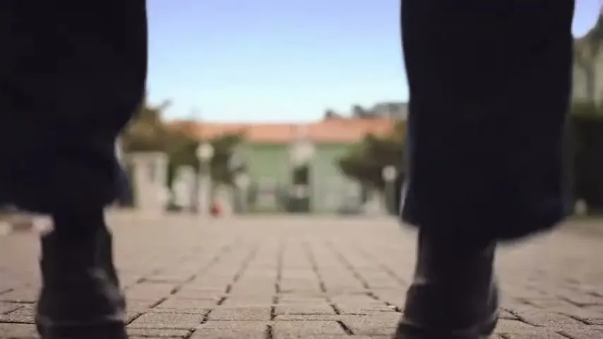 A person's legs and feet are seen from a low angle, walking on a paved path with blurred houses in the background.