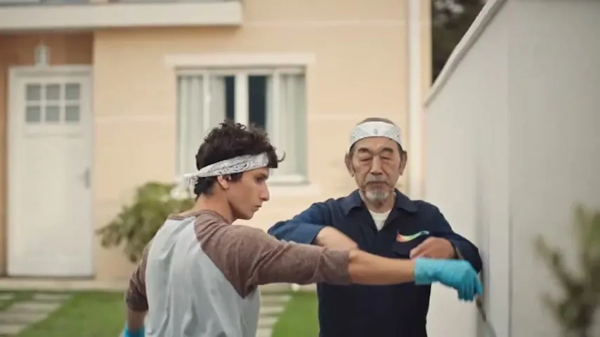 A young man in a bandana and an older man in a work uniform are painting a white wall outside a house, with the older man guiding the younger man's arm.